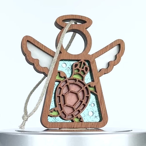 A wooden angel-shaped ornament with carved wings and a halo, featuring a sea turtle in the center. The turtle has a brown shell with green accents and is surrounded by a blue background with bubble-like patterns. A braided linen string is attached to the ornament.