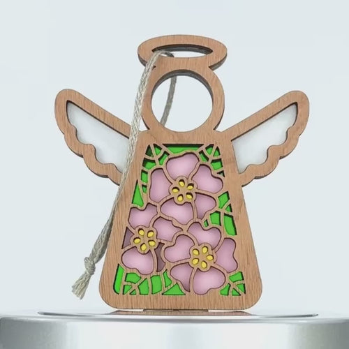 A wooden angel ornament with a hollow circular head, wings, and a halo. The body features a colorful stained glass–style design with pink evening primrose flowers and green leaves. A braided linen string is attached for hanging. The ornament stands against a plain white background.
