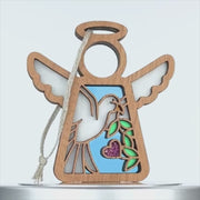 A wooden angel ornament with a cutout design. The angel has wings, a halo, and a string for hanging. Inside the body cutout, a dove ornament holding an olive branch and a small pink heart is visible against a blue background.