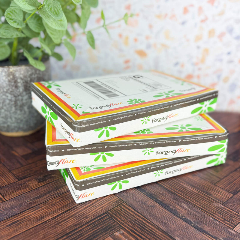 Two stacked Forged Flare® packages with floral designs are on a wooden surface, showcasing the brand's shipping protection worth $2.00 for purchases up to $300. A green plant is in the background against a subtle multicolored wall pattern.