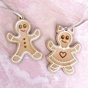 The Forged Flare® Sweet Gingerbread Cookies Ornaments: Boy & Girl Set (3.5"H) features a smiling boy with buttons and a girl with a dress and bow in white and pink, perfect for holiday decor against a light pink marbled background.