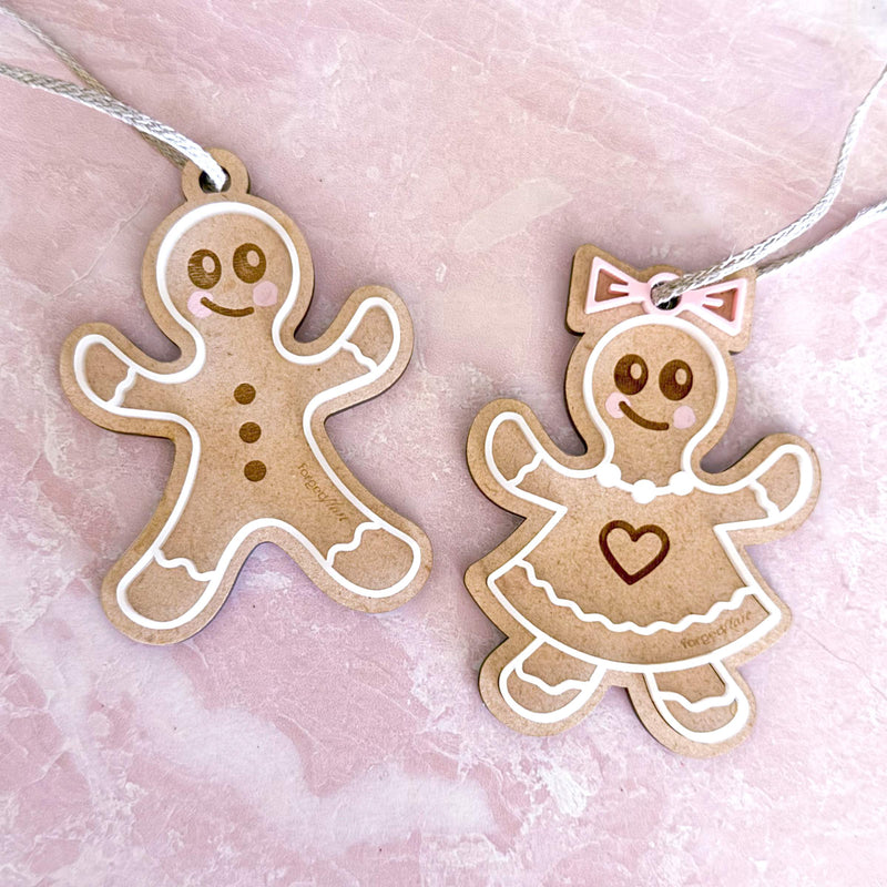 The Forged Flare® Sweet Gingerbread Cookies Ornaments: Boy & Girl Set (3.5"H) features a smiling boy with buttons and a girl with a dress and bow in white and pink, perfect for holiday decor against a light pink marbled background.