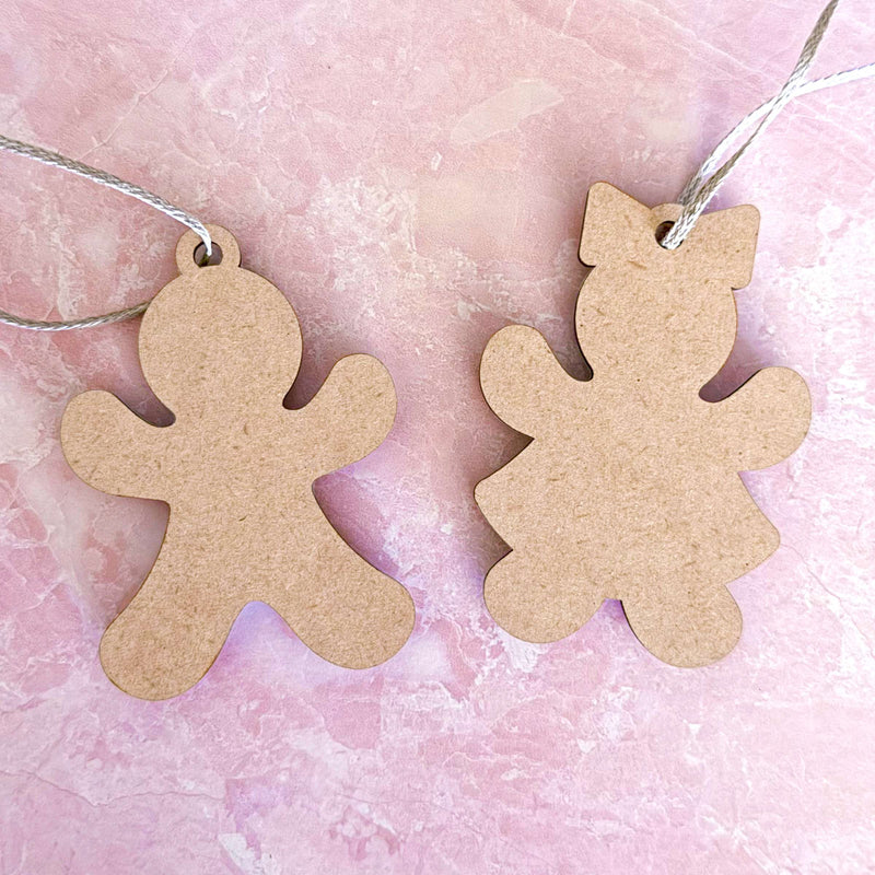 Two blank wooden Sweet Gingerbread Cookies Ornaments—a boy and a girl, each 3.5" tall—by Forged Flare® lie on a pink marbled surface. Each ornament comes with a string loop, perfect for hanging as festive holiday decor.