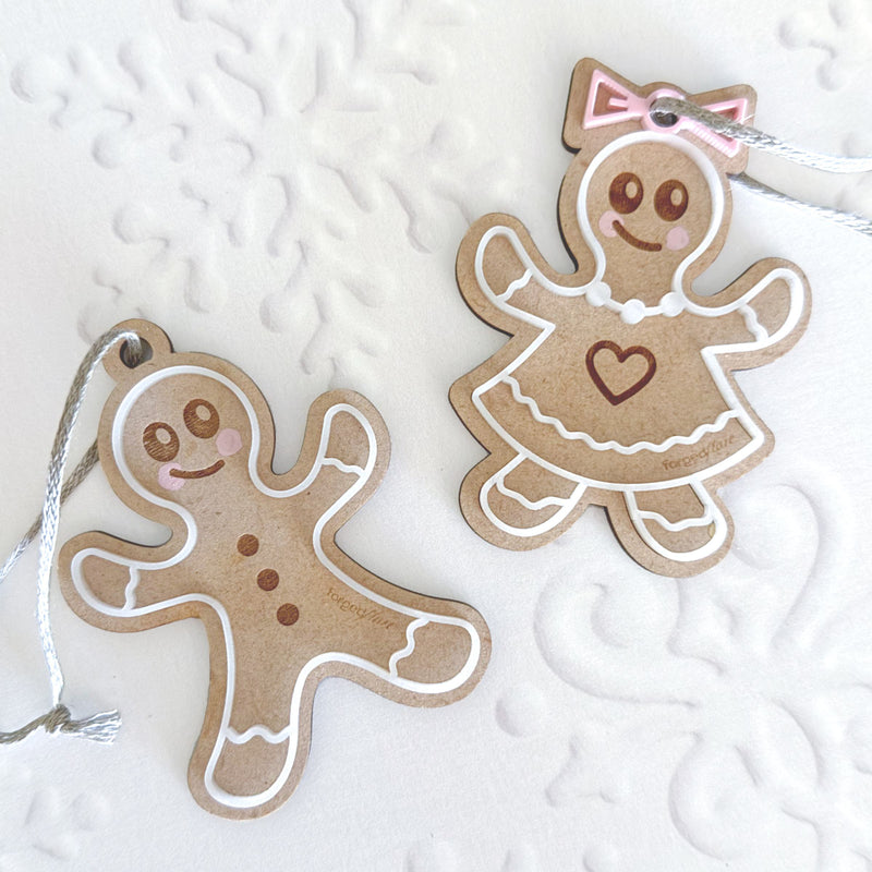 The Forged Flare® Sweet Gingerbread Cookies Ornaments: Boy & Girl Set, 3.5" (H), features a boy with buttons and a girl in a dress with heart and pink bow, both with silver hanging strings—perfect for charming holiday decor.