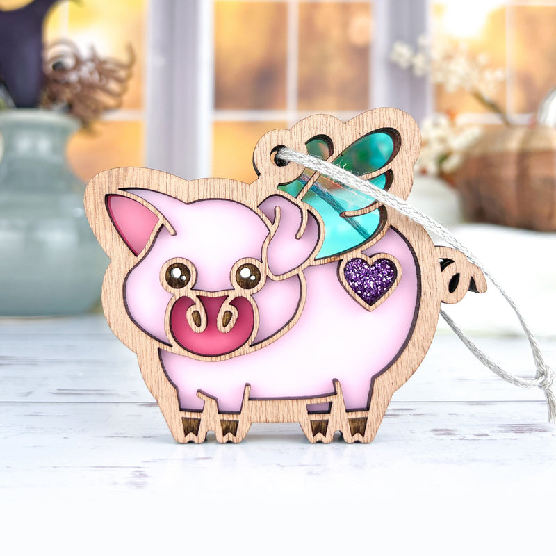 The Forged Flare® When Pigs Fly Ornament, 3" (H), features a pink pig with green wings and a purple glitter heart. With string attached, it's perfect for farmhouse mantel décor. Displayed on white with vases and a bright window in the background.