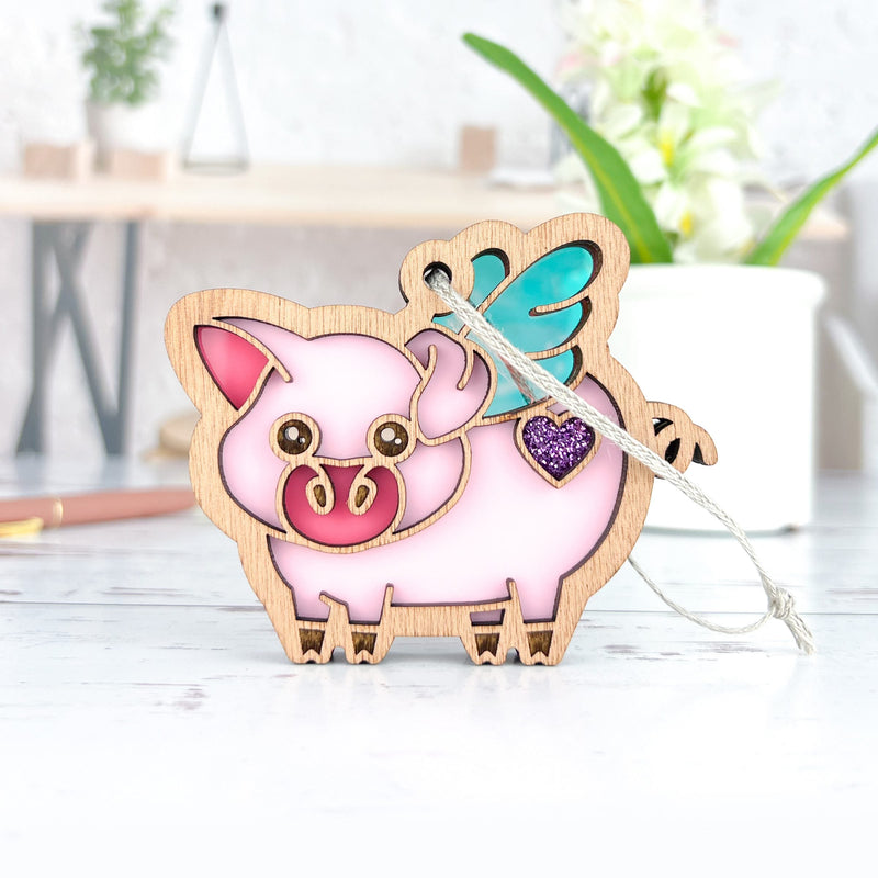 The Forged Flare® When Pigs Fly Ornament, 3" (H), is a cute wooden pig with teal wings and a purple glitter heart, hanging from a white string. Ideal for farm decor or bright farmhouse mantels, desks, or plant-filled rooms.