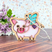 Add whimsical flair to your mantel with the Forged Flare® When Pigs Fly Ornament, a 3" pink wooden pig with blue wings and a purple glitter heart—perfect farmhouse decor nestled among purple flowers and green leaves.