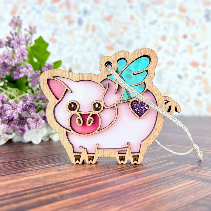 Add whimsical flair to your mantel with the Forged Flare® When Pigs Fly Ornament, a 3" pink wooden pig with blue wings and a purple glitter heart—perfect farmhouse decor nestled among purple flowers and green leaves.