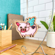The Forged Flare® When Pigs Fly Ornament, 3" (H), a pink pig with blue wings and a purple heart, hangs from a metal stand on a wooden desk, bringing whimsical farm charm to your office amidst supplies and a potted plant.