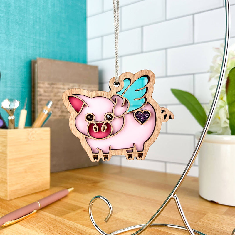 The Forged Flare® When Pigs Fly Ornament, 3" (H), a pink pig with blue wings and a purple heart, hangs from a metal stand on a wooden desk, bringing whimsical farm charm to your office amidst supplies and a potted plant.