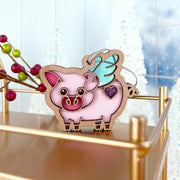The Forged Flare® When Pigs Fly Ornament, 3" (H), features a pink pig with teal wings and a purple heart standing on a gold shelf—ideal for farmhouse mantels. Snowy trees blur behind, and red berries accent the left side.