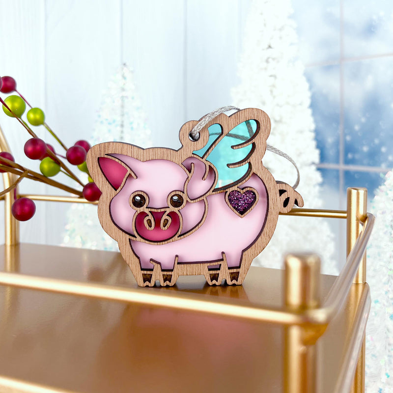 The Forged Flare® When Pigs Fly Ornament, 3" (H), features a pink pig with teal wings and a purple heart standing on a gold shelf—ideal for farmhouse mantels. Snowy trees blur behind, and red berries accent the left side.
