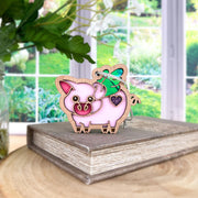 The Forged Flare® When Pigs Fly Ornament, 3" (H), is a cute pink pig with a heart and green bow, perched on a closed book—perfect farmhouse mantel decor. Set on wood with a blurred garden visible through a large window.