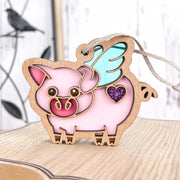 The Forged Flare® When Pigs Fly Ornament, 3" (H), features a pink pig with blue wings and a purple glitter heart—ideal as a whimsical farmhouse mantel decor piece. The smiling ornament adds charm wherever it's displayed.