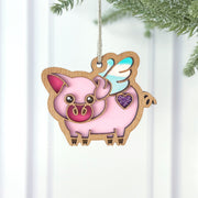 The Forged Flare® When Pigs Fly Ornament, 3" (H), features a pink pig with blue wings and a purple glitter heart, outlined in gold and smiling—perfect for adding whimsical charm to your farmhouse mantel or farm decor.
