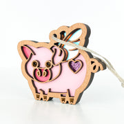 The Forged Flare® When Pigs Fly Ornament, 3" (H), features pink accents, a glittery purple heart, and a multicolored tail. Perfect whimsical farm decor or mantel piece. Includes string for easy hanging against a white background.
