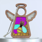A wooden angel ornament with outstretched wings. The body is filled with a colorful stained glass–style design featuring a honey bee with blue wings and a yellow and black body, against a purple background with yellow hexagon shapes. A string is attached to the top for hanging.