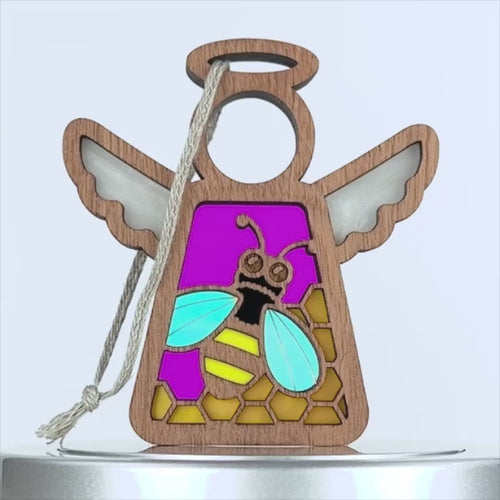 A wooden angel ornament with outstretched wings. The body is filled with a colorful stained glass–style design featuring a honey bee with blue wings and a yellow and black body, against a purple background with yellow hexagon shapes. A string is attached to the top for hanging.