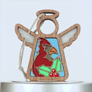 A wooden angel ornament with wings and a halo, featuring a colorful cardinal bird in the center. The charming cardinal ornament is holding green holly leaves with red berries at the bottom. A string is attached at the top for hanging. The background is white.