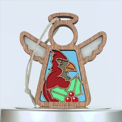 A wooden angel ornament with wings and a halo, featuring a colorful cardinal bird in the center. The charming cardinal ornament is holding green holly leaves with red berries at the bottom. A string is attached at the top for hanging. The background is white.