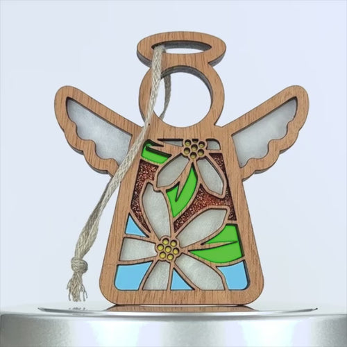 Wooden angel decoration with a stained glass–style orange blossom. The angel has cut-out wings, a halo, and an orange blossom floral design featuring white, green, sparkly orange, and blue colors. A piece of braided linen string is looped through a hole near the wing for hanging.