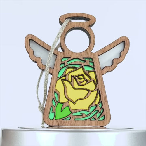 A wooden angel decoration with a stained glass–style design featuring a yellow rose ornament in the center. The angel has outstretched wings and a halo, with a braided linen string for hanging. The overall design combines elements of natural wood and colorful pieces.