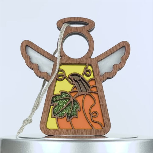 A wooden angel ornament with carved wings, a halo, and a rope loop for hanging. The body features a colorful design with a pumpkin, leaves, and a vibrant yellow-orange background. This charming pumpkin ornament is set against a plain white backdrop.