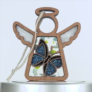 A wooden angel ornament with transparent wings and body. This charming blue butterfly ornament features a glittery butterfly in the center and has a braided linen string attached for hanging. Background is plain white.