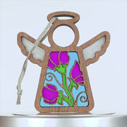 A wooden angel sweet pea ornament with a halo, featuring stained glass-style panels colored in purple, green, and blue. The angel has a cutout pattern with floral designs on its body. A braided linen string is attached at the top for hanging. The background is white.