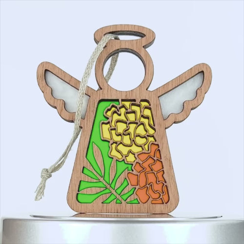 A marigold ornament crafted from wood features cut-out wings and a halo. The body showcases vibrant stained glass-style sections displaying yellow and orange flowers with green leaves. A braided linen string is attached for hanging against a plain white background.