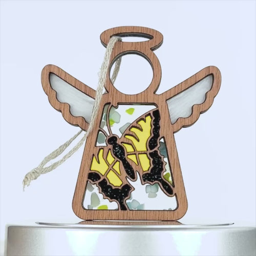 A wooden angel ornament with outstretched wings and a halo has a decorative design featuring two yellow swallowtail butterflies. A braided linen string is attached to the top for hanging. The background is plain white.