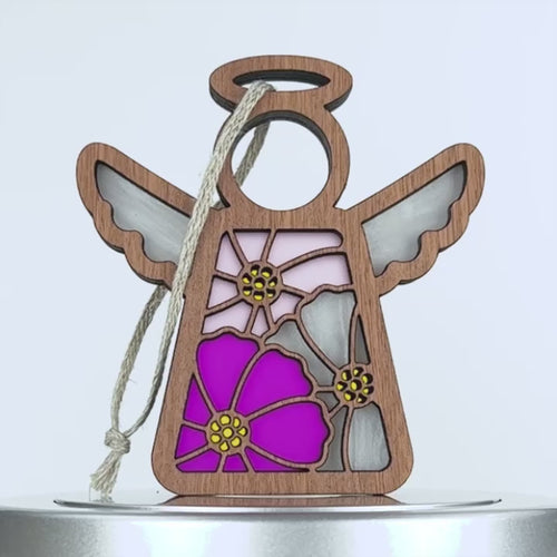 A decorative wooden angel with a colorful stained glass effect in the shape of cosmos flowers fills the body. The angel has outstretched wings and a halo, with a piece of braided linen string attached for hanging. The colors in the stained glass are pink, magenta, and yellow accents.
