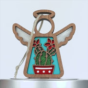 A wooden angel ornament with white wings and a halo. In the middle, there's a turquoise background showcasing a charming cactus design in a red pot with red flowers. A braided linen string is attached to the top for easy hanging.