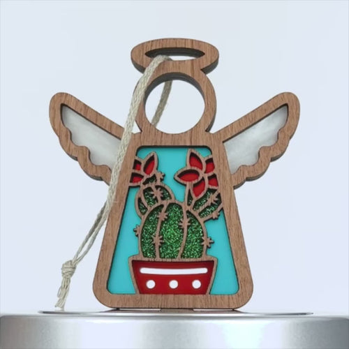 A wooden angel ornament with white wings and a halo. In the middle, there's a turquoise background showcasing a charming cactus design in a red pot with red flowers. A braided linen string is attached to the top for easy hanging.