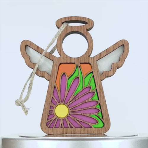 A wooden angel ornament with outstretched wings featuring a colorful design. The angel has a bright purple flower with a yellow center on its body and intricate patterns on the wings. There is a loop of twine attached to the top for hanging.