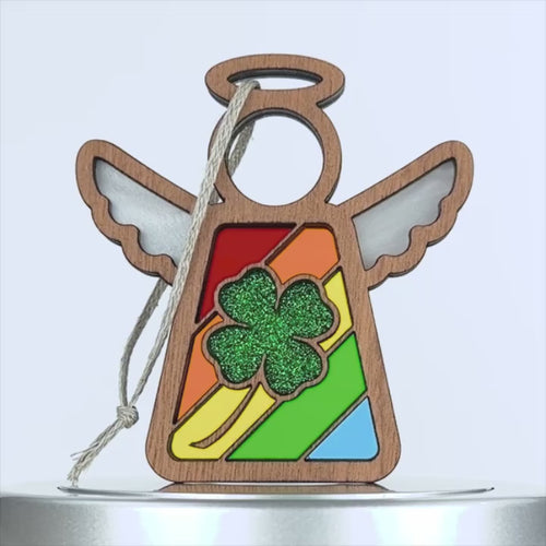 A wooden angel ornament with white wings, a halo, and a colorful stained glass–style body featuring a green glittery four leaf clover in the center. A braided linen string hangs from the top left for easy display. The background is plain white.