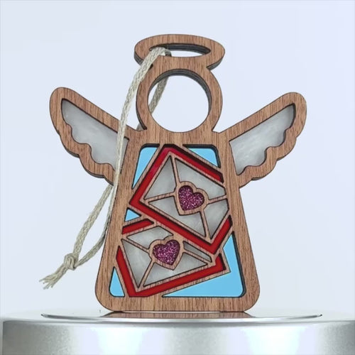 A wooden angel-shaped ornament with a halo and wings, perfect as a Valentine's Day ornament. The body showcases two envelopes adorned with red hearts, while the wings and envelopes sparkle with glitter detailing. A braided linen string is attached at the top for hanging against a plain white background.