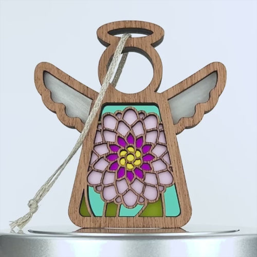 A wooden Christmas ornament shaped like an angel with a halo, featuring colorful stained glass–style wings and a vibrant pink and yellow chrysanthemum flower design on its body. It has a braided linen string attached for hanging.