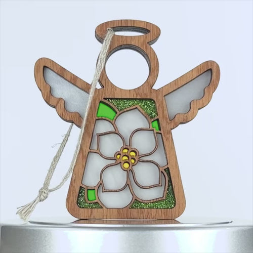 A wooden angel ornament with a stained glass–style design features a white magnolia flower with yellow and green accents. The angel has a halo and wings, and a string loop for hanging at the top. The background is plain white.