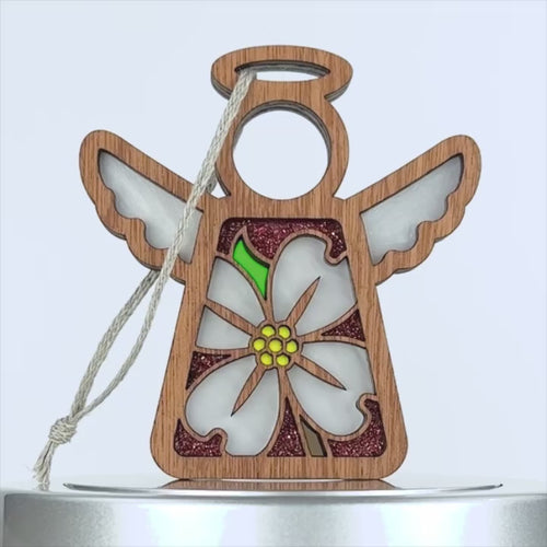 A wooden angel ornament with cut-out wings and a lovely floral design featuring a delicate dogwood flower. The blossom has white petals with a yellow center and a green leaf. A string is attached to the top for hanging against the plain white background.