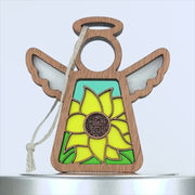 A sunflower ornament featuring a wooden angel decoration with stained glass–style elements. The angel's body showcases a vibrant yellow sunflower against a light blue background. The decoration includes a braided linen string for hanging.
