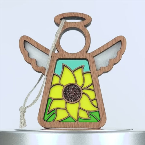 A sunflower ornament featuring a wooden angel decoration with stained glass–style elements. The angel's body showcases a vibrant yellow sunflower against a light blue background. The decoration includes a braided linen string for hanging.
