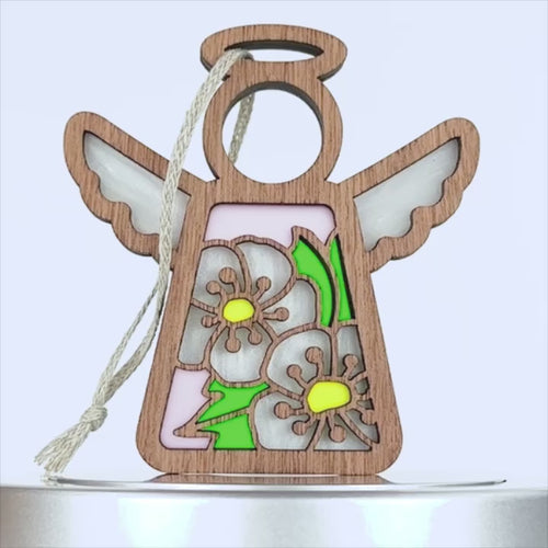 A wooden angel hawthorn ornament with a cutout design features white flowers with yellow centers and green leaves set against a pink background. It has a braided linen string attached for hanging. The angel's wings and halo are distinctly shaped, enhancing its decorative appeal.