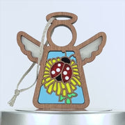 A wooden angel ornament with a string loop for hanging, featuring translucent wings and a central design of a red ladybug on a vibrant dandelion flower. The background is partially filled with light blue, enhancing the sstained glass–style effect.