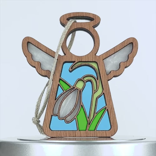 A wooden angel snowdrop ornament with transparent wings and a halo at the top. The body features a stained glass–style design with a white snowdrop flower and green leaves set against a light blue background. The decoration is accompanied by a braided linen string.