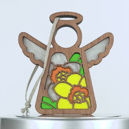 A wooden angel ornament with a stained glass–style design featuring white and yellow daffodil flowers and green leaves. The angel has white stained glass–style wings, a halo, and a braided linen string for hanging. The background is plain white.