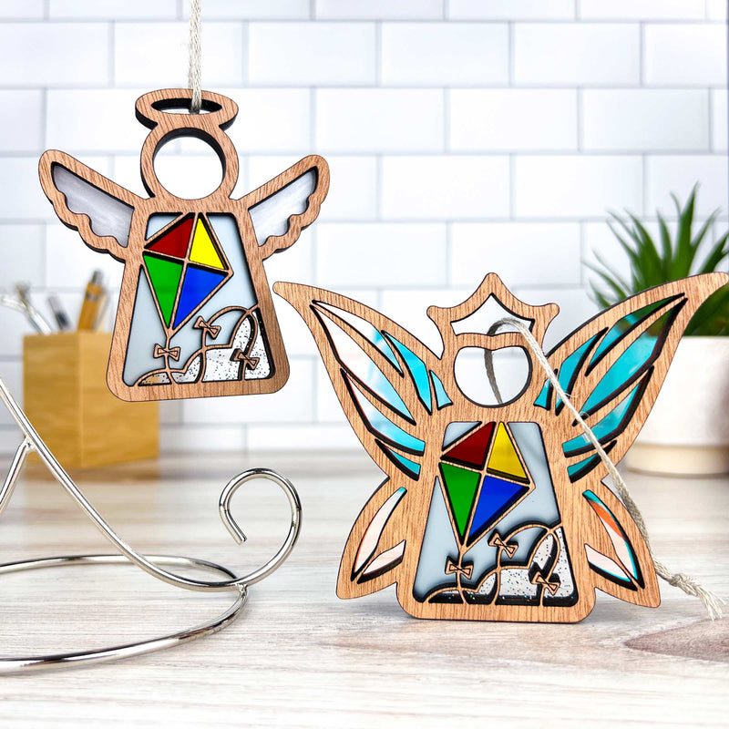 Two Forged Flare® Fairy - Fly A Kite for Charity Ornaments, 3.7" tall with stained glass-style wings and colorful geometric shapes, are displayed on a light wooden surface with a white tile background and a small potted plant.