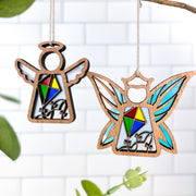 Two Forged Flare® "Fairy - Fly A Kite for Charity" ornaments, each 3.7" high with stained-glass-style designs and "LA" letters, hang side by side against white tiles, creating unique fairy decor and thoughtful gifts for women. Green leaves peek in the foreground.