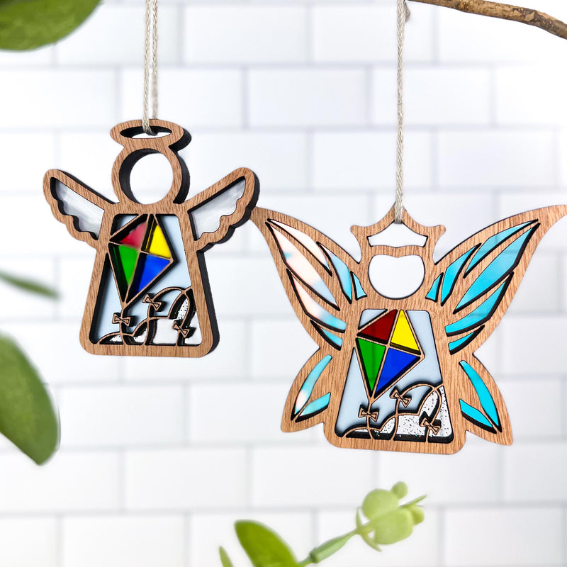 Two Forged Flare® "Fairy - Fly A Kite for Charity" ornaments, each 3.7" high with stained-glass-style designs and "LA" letters, hang side by side against white tiles, creating unique fairy decor and thoughtful gifts for women. Green leaves peek in the foreground.