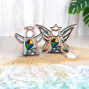 Two Forged Flare® Fly A Kite for Charity Mother's Angels® 3.5" wooden ornaments, featuring colorful kite designs, stand on sand near a seashell—ideal for beach house decor—with a leafy plant and light wood backdrop behind them.